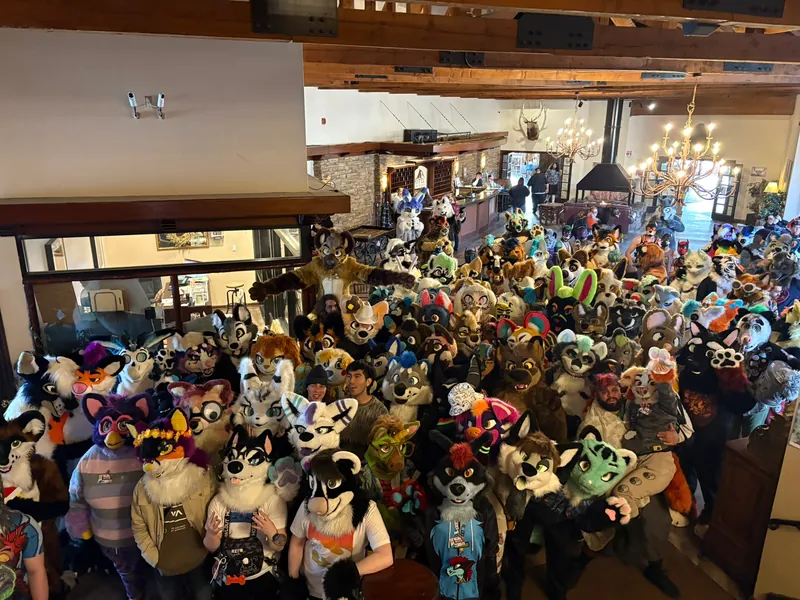 The 2025 group photo: a lodge lobby packed with fursuiters under wooden beams and chandeliers, seen from the balcony