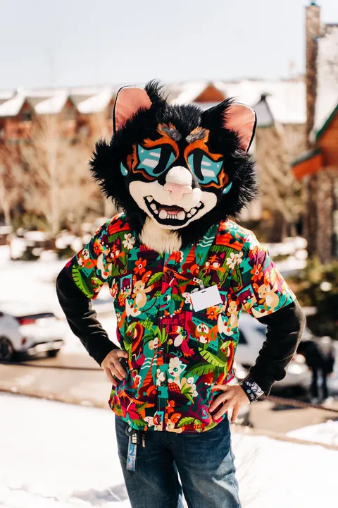 A fursuiter with a black cat head and a bright tropical print shirt standing in the snow